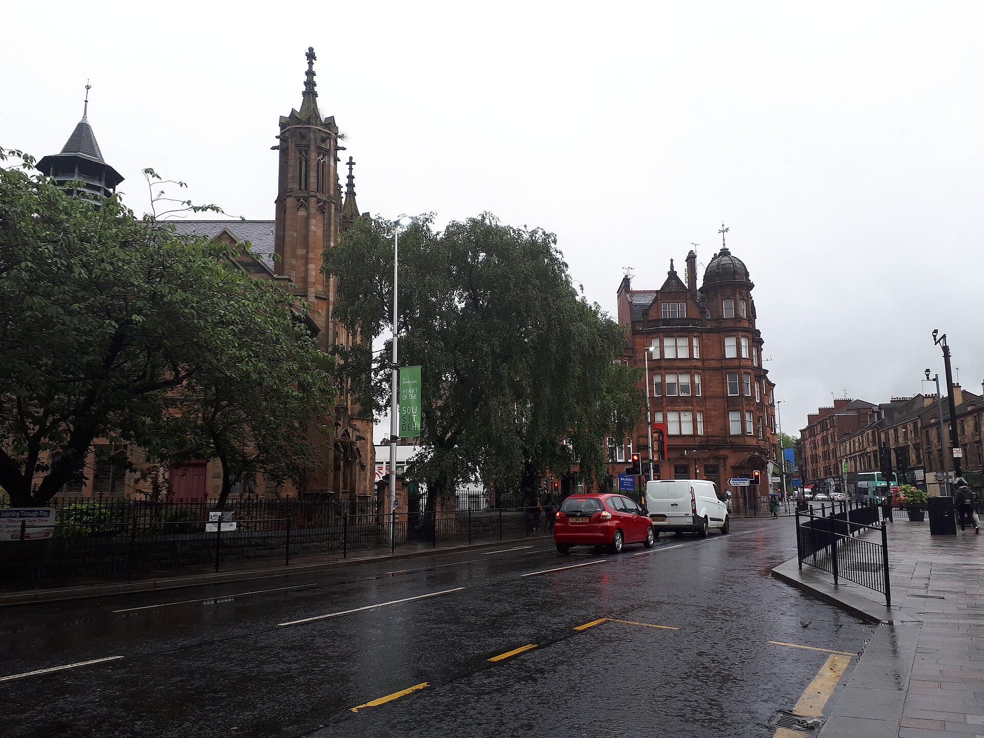 A wet main road through Shawlands with a Gothic church spire among trees on the left, a red sandstone tenement corner with a domed turret ahead, and cars and vans along the street