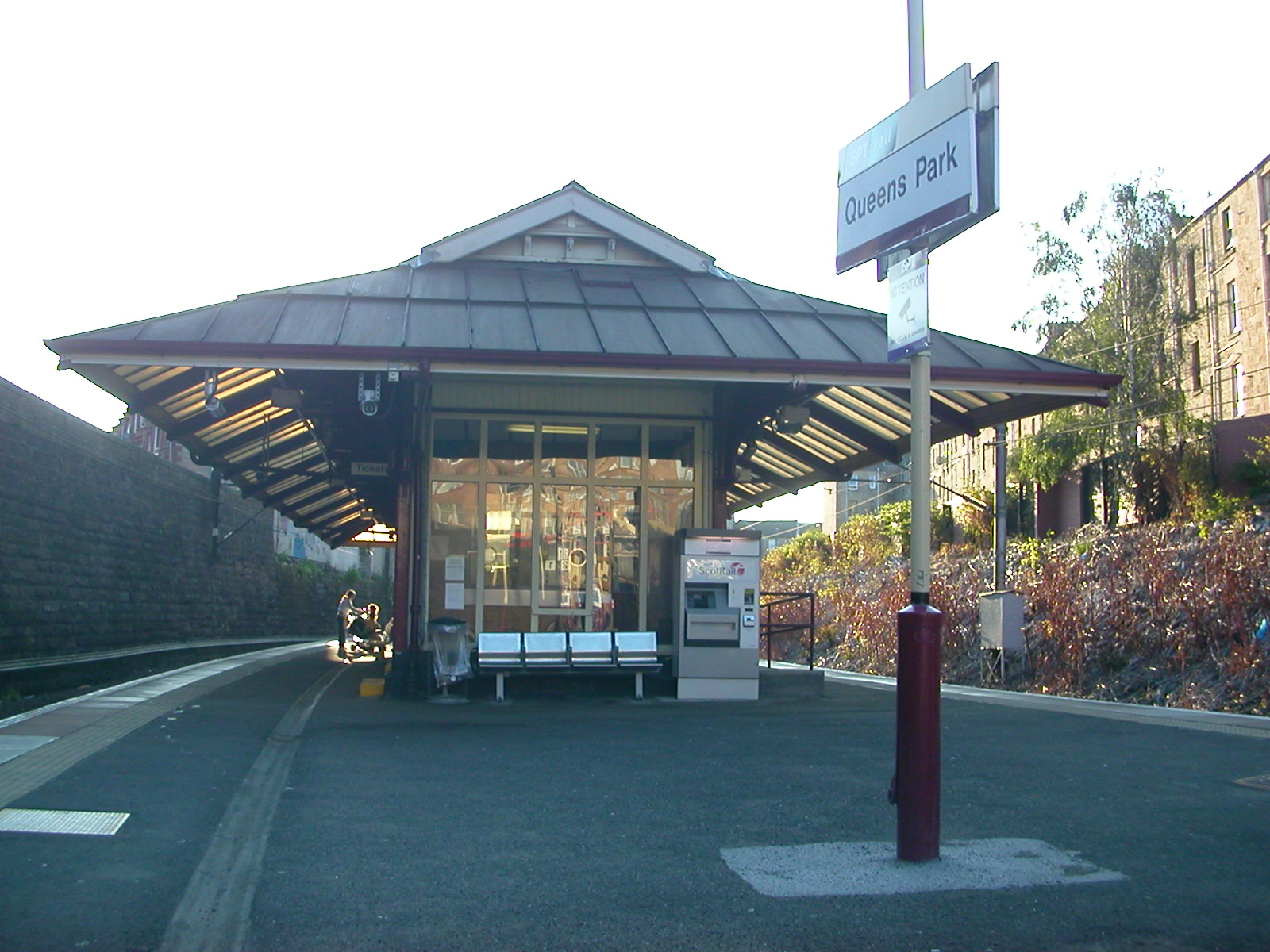 The platform at Queen's Park station in Glasgow, with a small pavilion-style station building, a bench and a Queens Park sign on a sunny day
