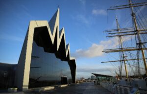 The zigzag roofline and glass end wall of the Riverside Museum on the Clyde quayside, with the masts and rigging of the tall ship Glenlee alongside under a blue sky