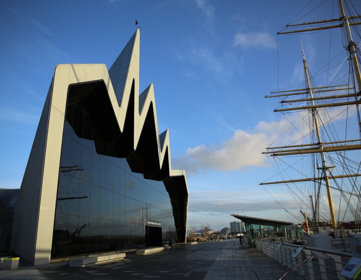 The zigzag roofline and glass end wall of the Riverside Museum on the Clyde quayside, with the masts and rigging of the tall ship Glenlee alongside under a blue sky