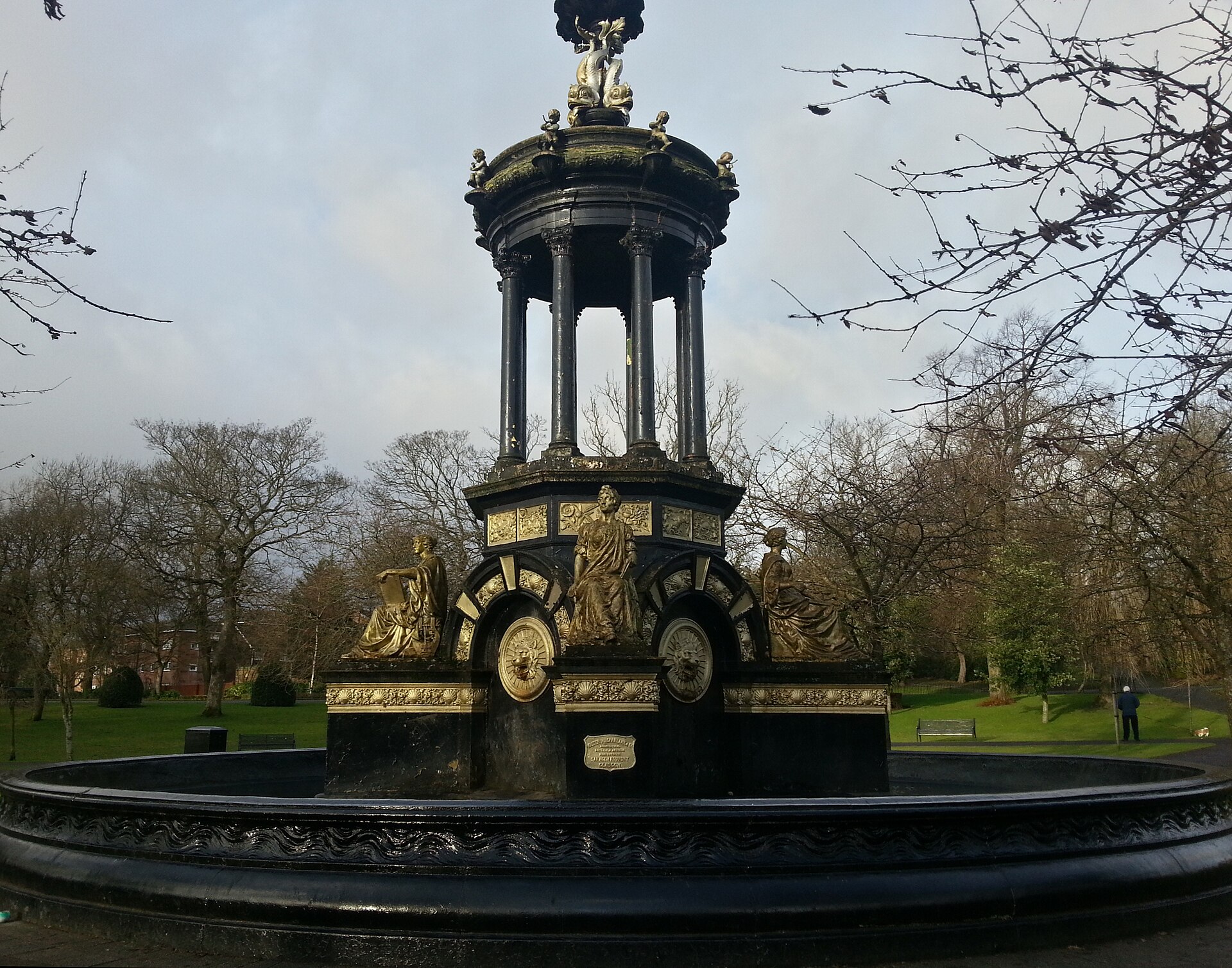 An ornate black and gold cast-iron fountain with a domed canopy on slender columns and seated figures around its base, in a circular basin in a park with bare winter trees