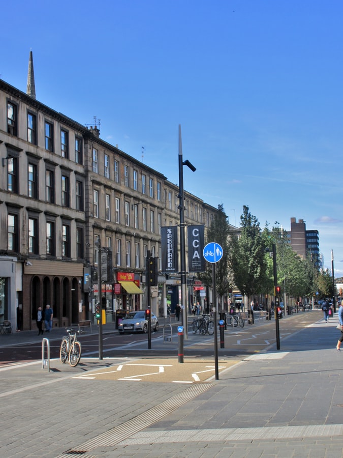 The western stretch of Sauchiehall Street on a sunny day, with sandstone terraces on the left, young street trees, wide paved pavements, a cycle lane and bicycles parked by a junction