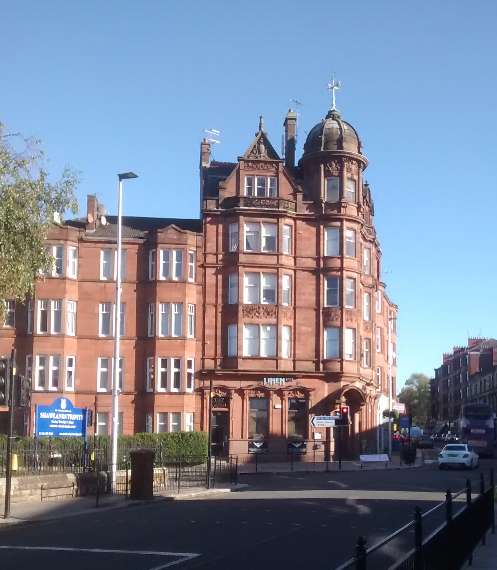 A tall red sandstone corner building at Shawlands Cross with bay windows, carved stonework and a domed corner turret, under a clear blue sky