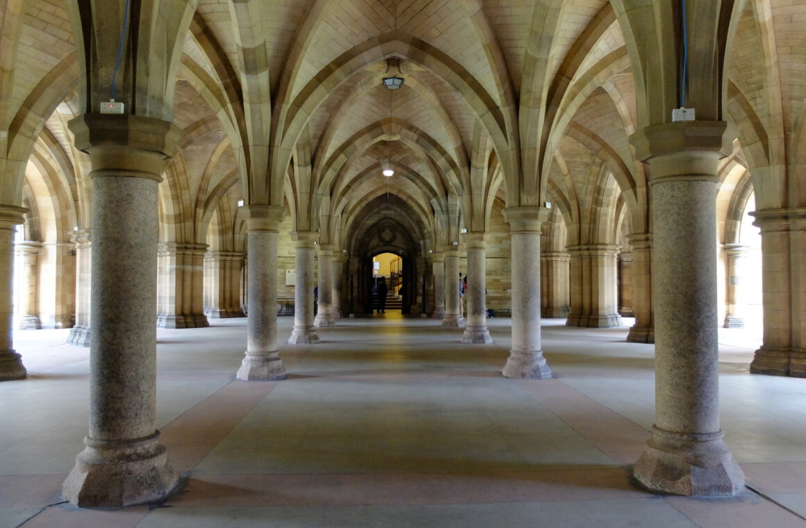 The vaulted sandstone cloisters beneath the University of Glasgow, with rows of stout columns and ribbed arches leading to a lit doorway at the far end