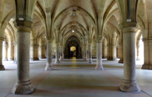 The vaulted sandstone cloisters beneath the University of Glasgow, with rows of stout columns and ribbed arches leading to a lit doorway at the far end