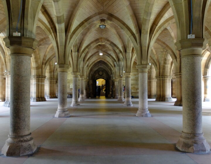 The vaulted sandstone cloisters beneath the University of Glasgow, with rows of stout columns and ribbed arches leading to a lit doorway at the far end