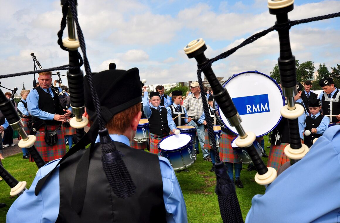 Seen over a piper's shoulder, a young pipe band in pale blue shirts and tartan kilts warms up on the grass at the World Pipe Band Championships, with a bass drum marked RMM