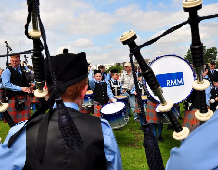 Seen over a piper's shoulder, a young pipe band in pale blue shirts and tartan kilts warms up on the grass at the World Pipe Band Championships, with a bass drum marked RMM
